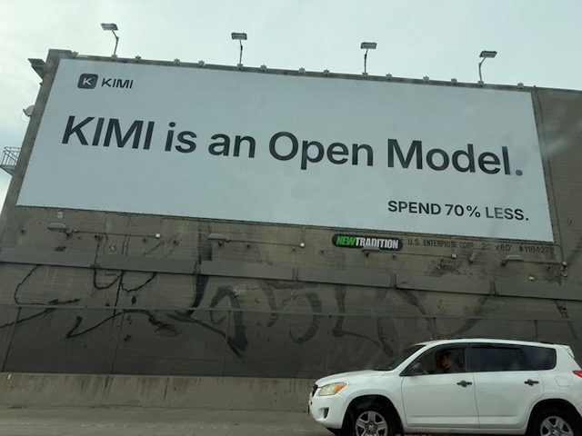 Billboarding reading “KIMI is an Open Model.” in large centered text, with a small lower right caption reading “Spend 70% less.”