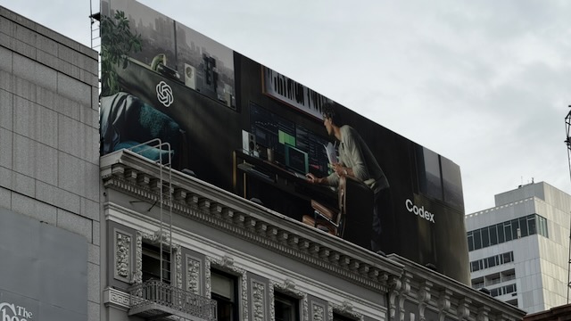 A billboard showing a person holding a takeout Chinese food container, while bending over their clearly very expensive home office, to do something on the laptop. The only caption is the OpenAI logo and the text “Codex”.